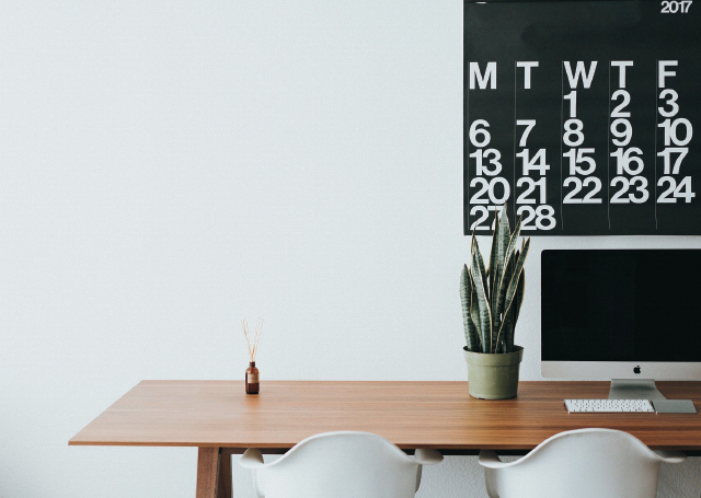 A desk with a plant and monitor with 2 chairs and a calendar on the wall.